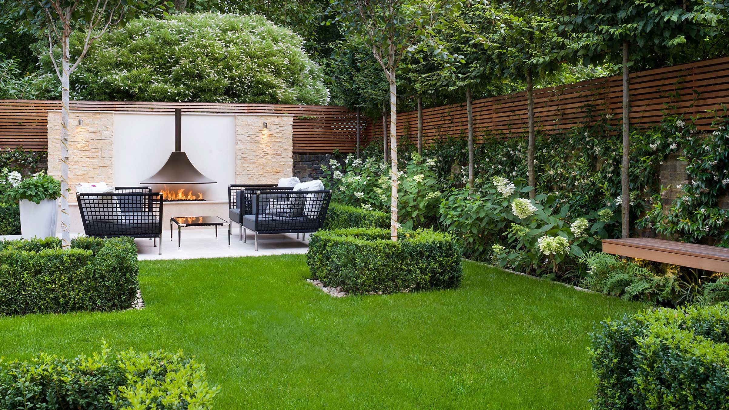 A backyard lounge area with a low fire pit, cushioned seating, and soft landscape lighting illuminating shrubs and trees at dusk