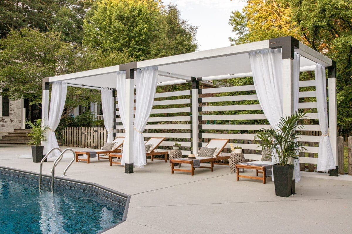 A backyard pergola with white outdoor curtains tied back, a cushioned daybed, and potted olive trees on a stone patio in warm afternoon light