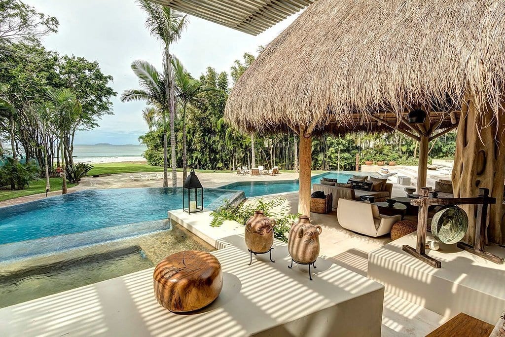 A beachfront villa in Punta Mita framed by palm trees, with lounge chairs on a stone terrace and calm ocean water beyond