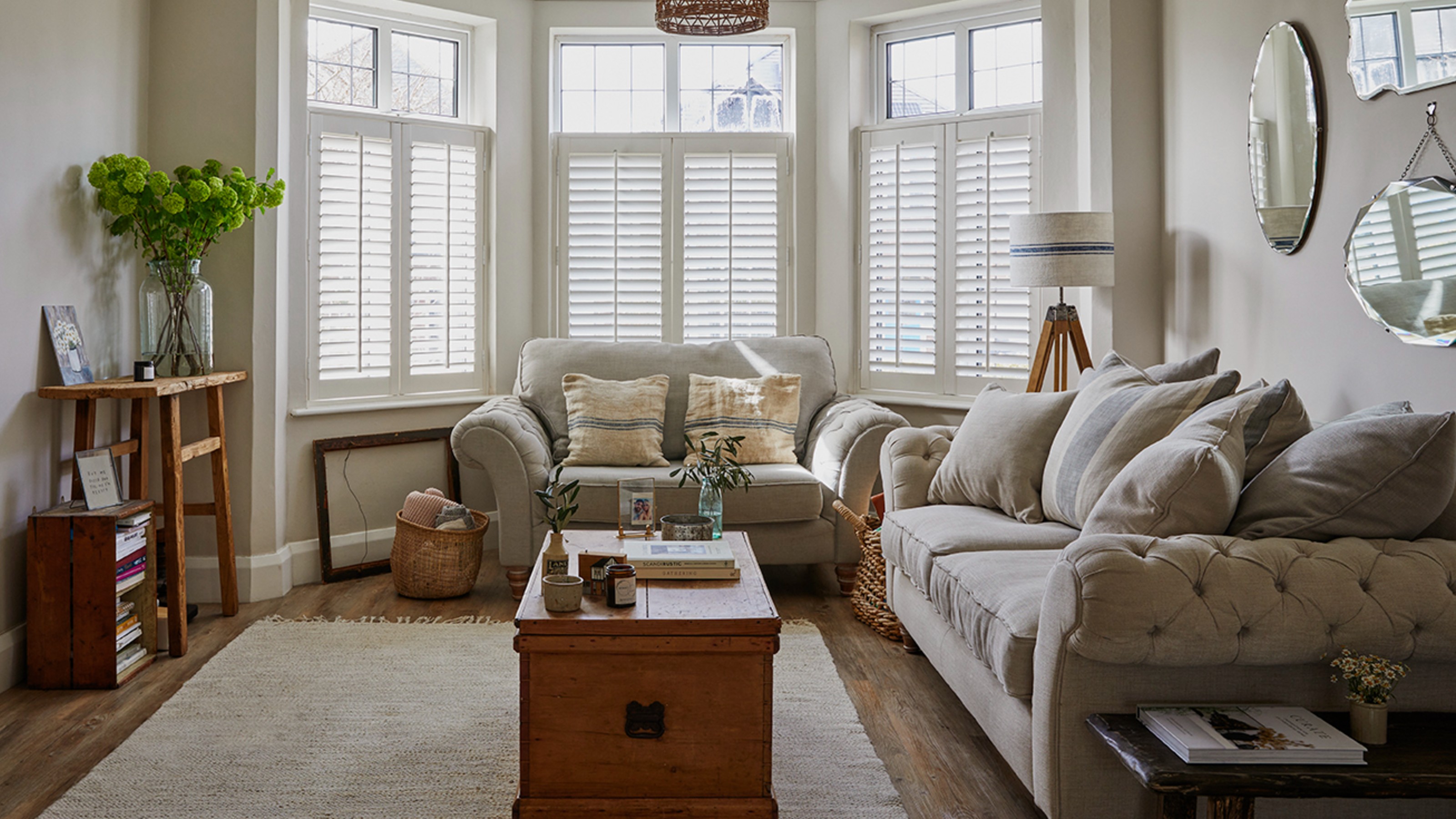 A bright, neutral-toned living room with a large sofa, layered throw pillows, a textured rug, and a sculptural coffee table, styled in a calm, celebrity-home-tour look