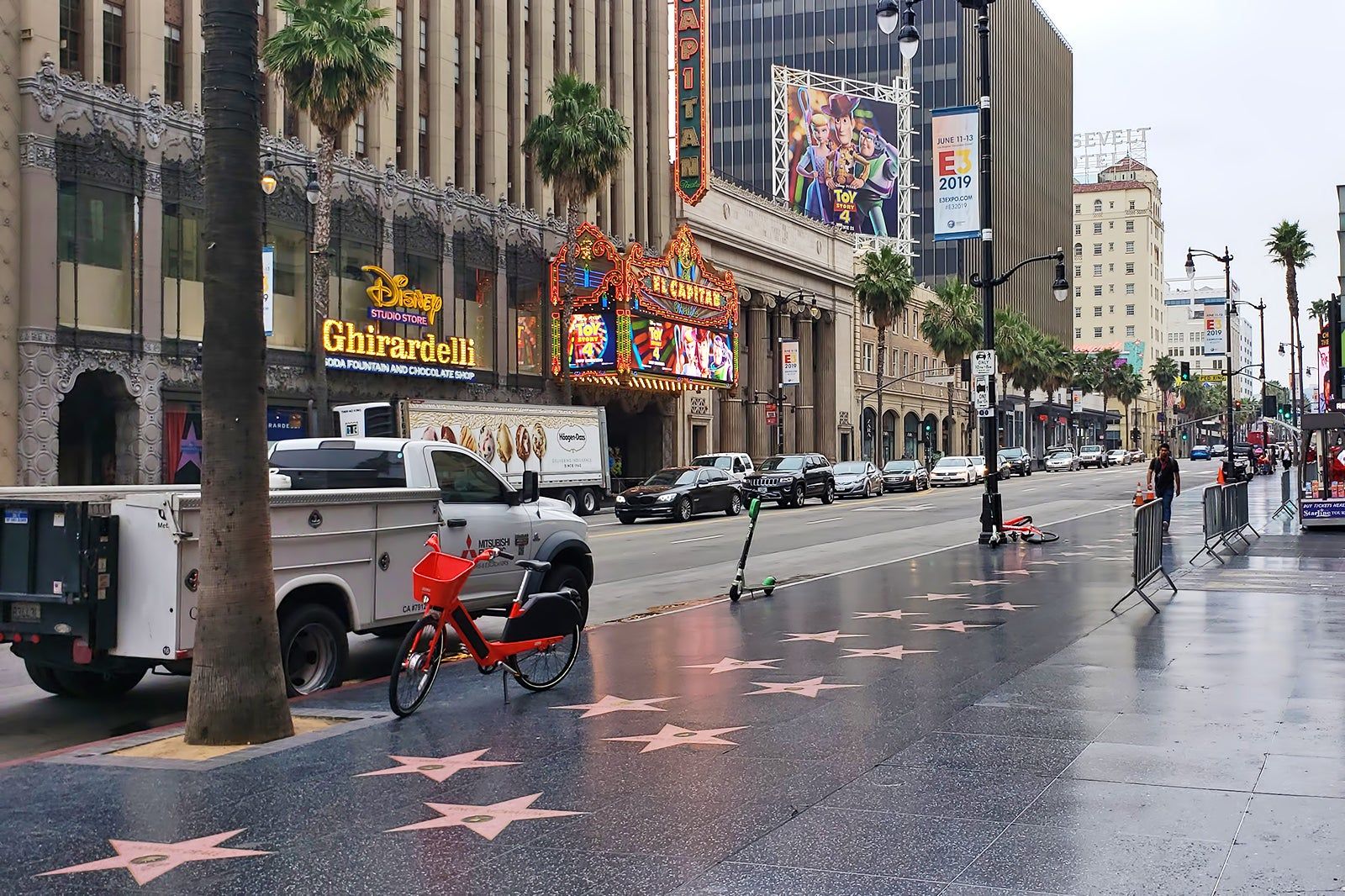A busy daytime view of Hollywood Boulevard with multiple Walk of Fame stars visible on the sidewalk and pedestrians walking past storefronts