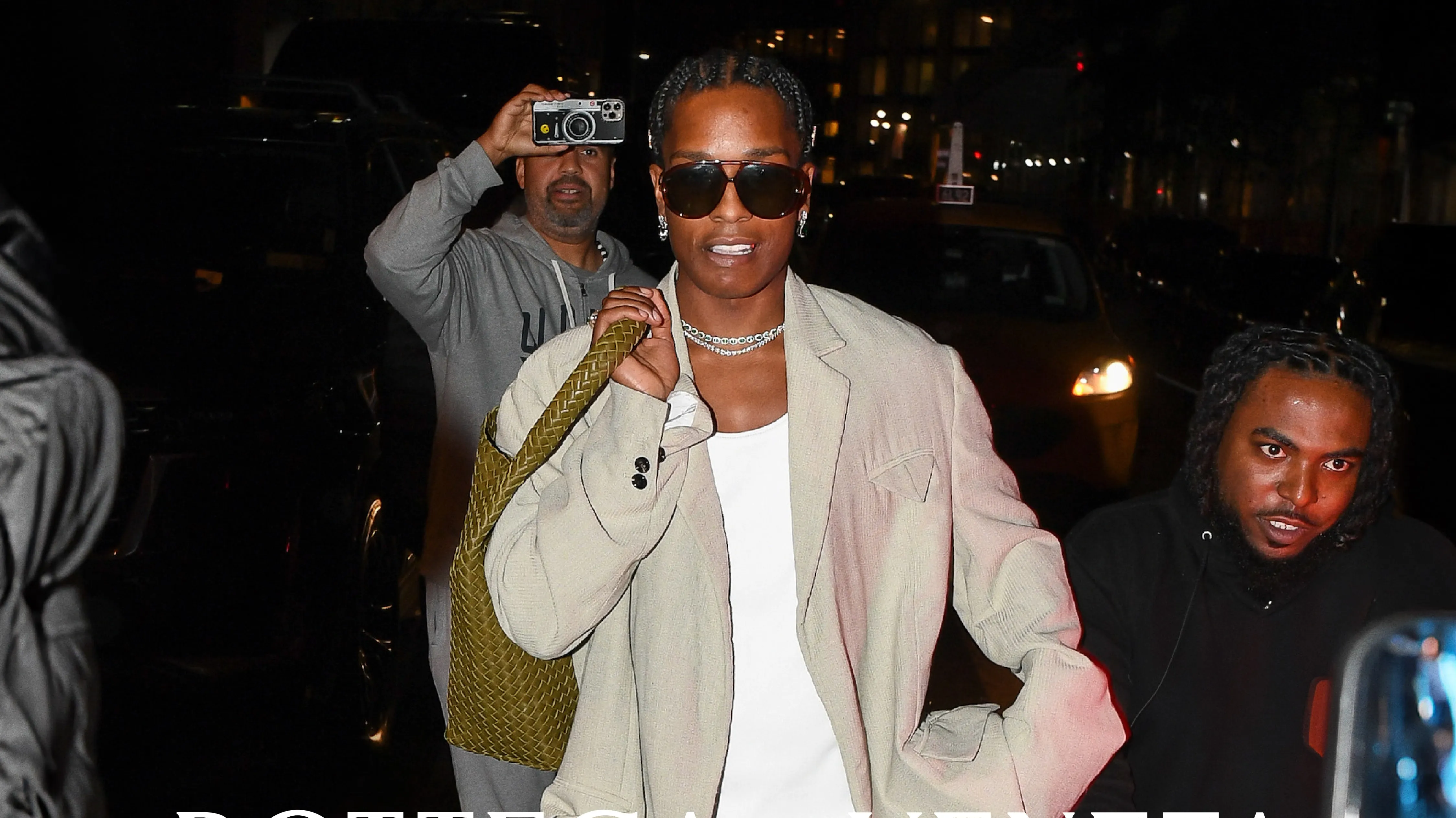 A celebrity on a city sidewalk adjusting their sunglasses while walking calmly as a photographer captures the moment from across the street