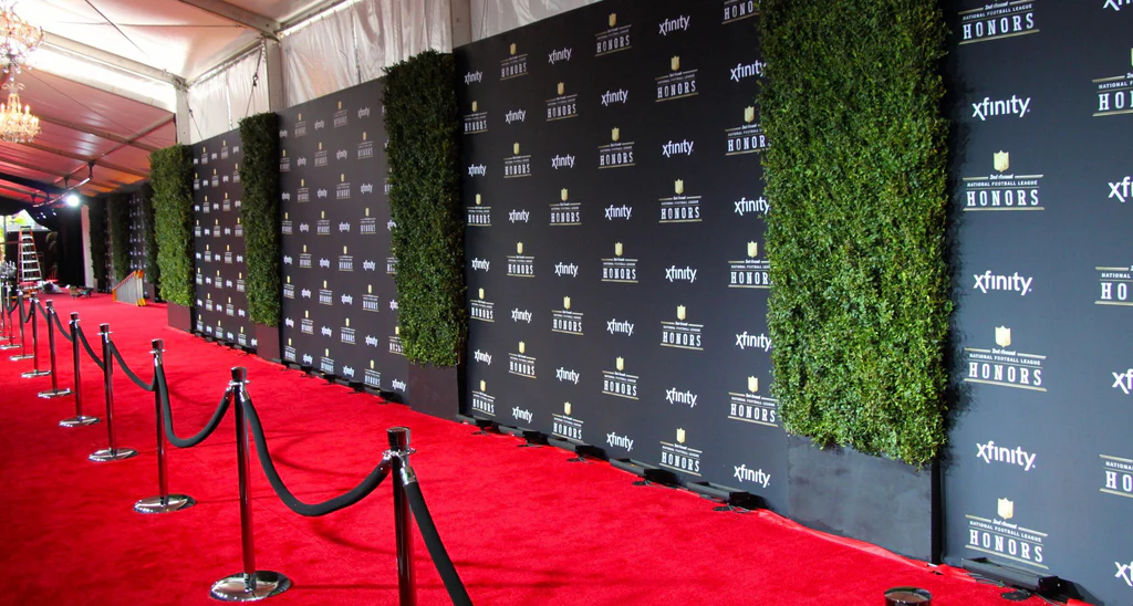 A celebrity posing on a red carpet in front of a step-and-repeat backdrop with repeated logos, with photographers visible behind the barricade
