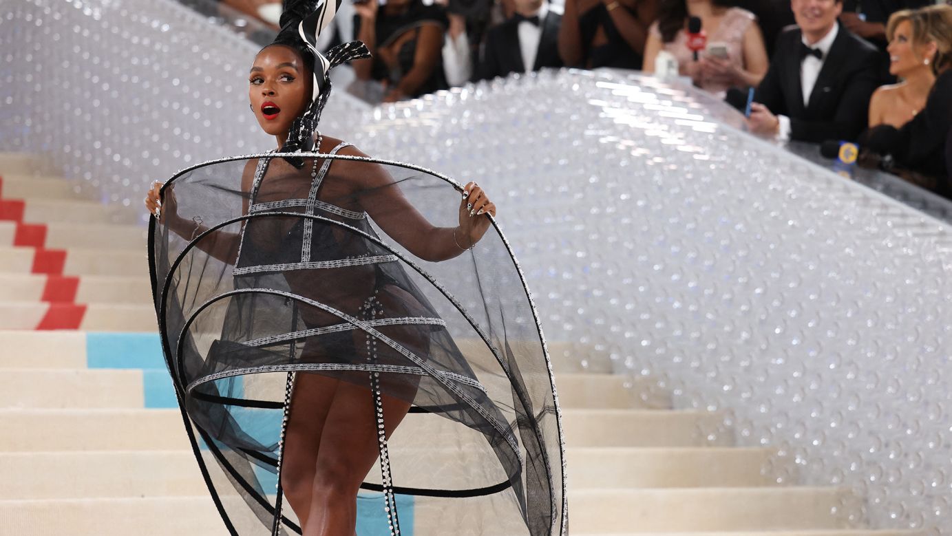 A celebrity posing on the Met Gala steps wearing an ornate couture gown with a dramatic cape and an embellished headpiece, photographed by a crowd of paparazzi