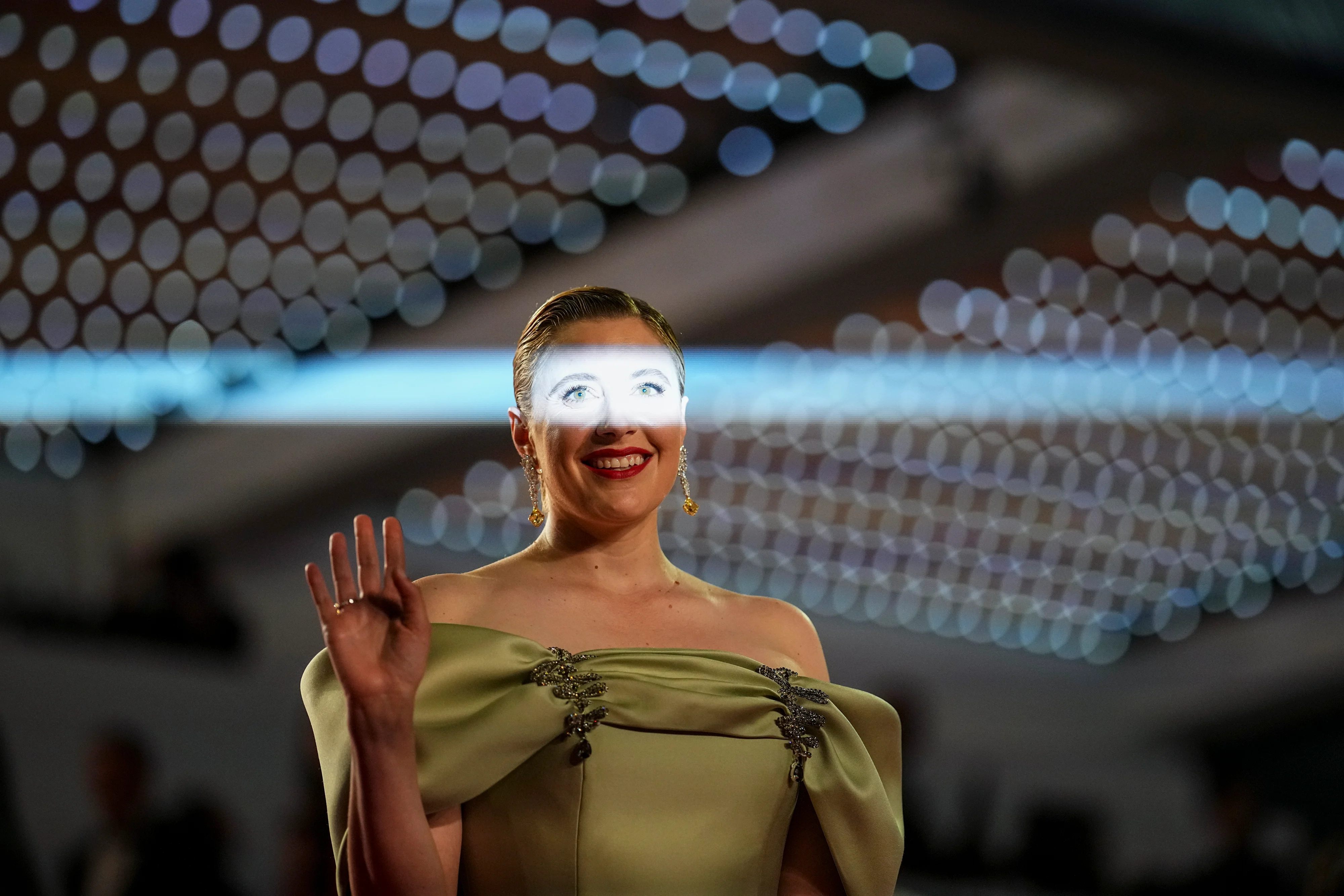 A celebrity smiling at a media wall event under flash lighting, with makeup visible and natural skin texture still present