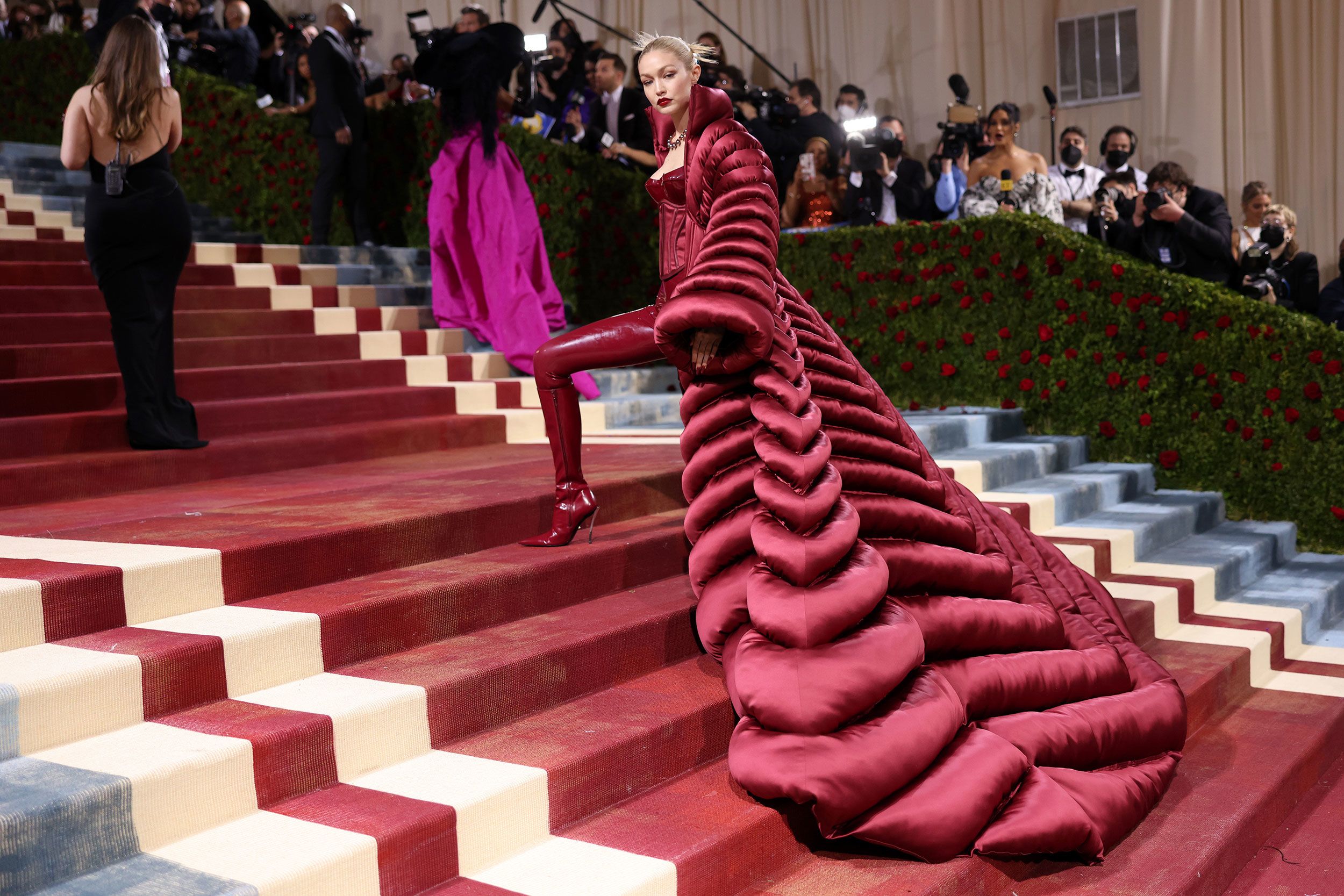 A celebrity standing on the Met Gala staircase at night in New York wearing a highly structured couture look with ornate detailing, photographed as flashes light up the scene