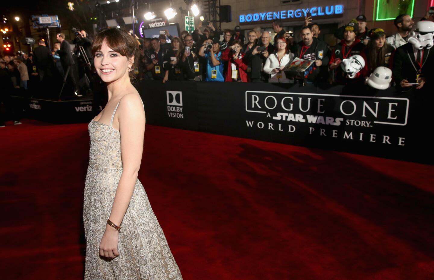 A celebrity walking a Hollywood premiere red carpet at night with photographers and fans behind barricades