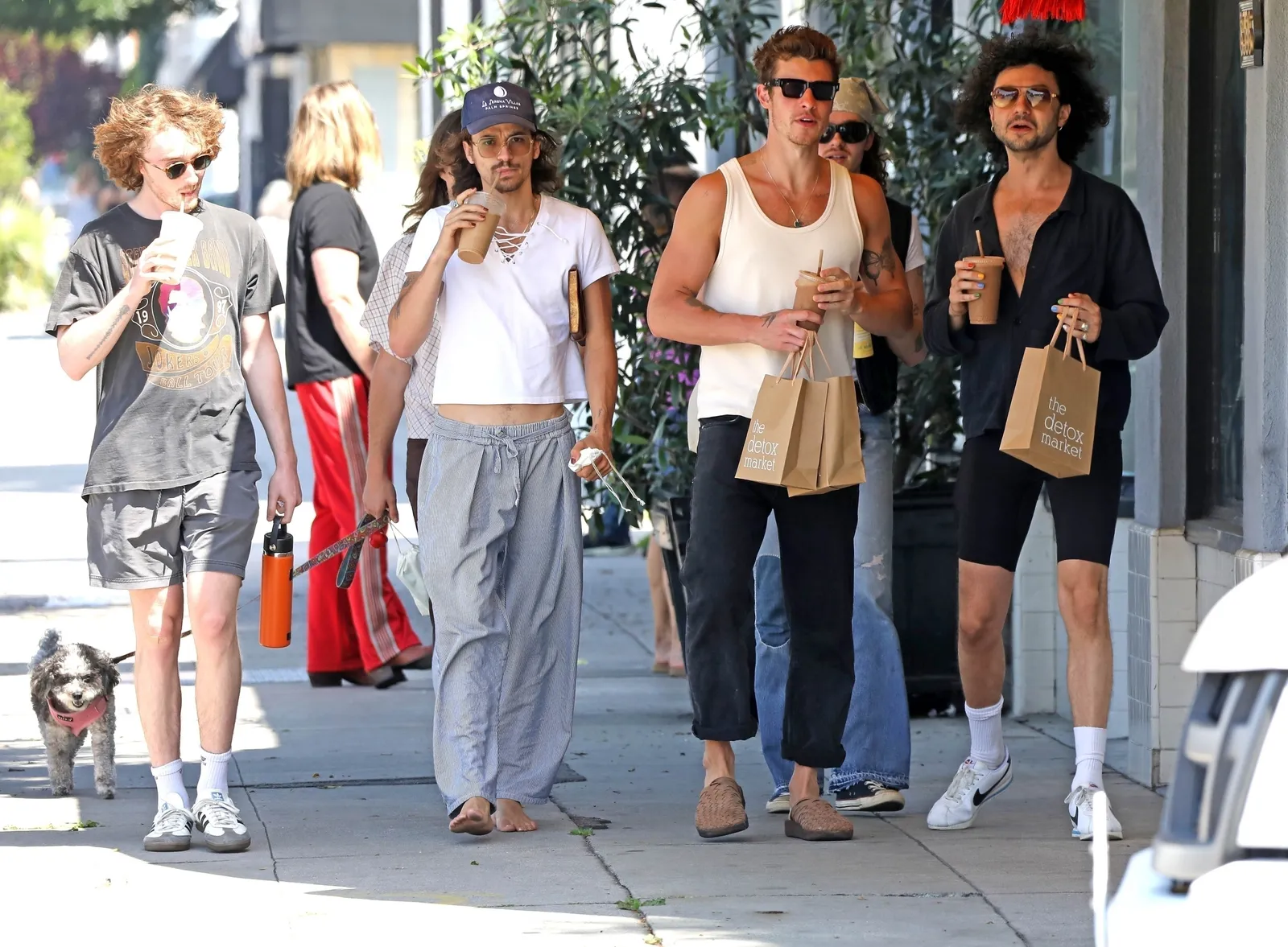 A celebrity walking past parked cars on a Los Angeles street with a relaxed expression while a photographer captures full-body shots from the curb