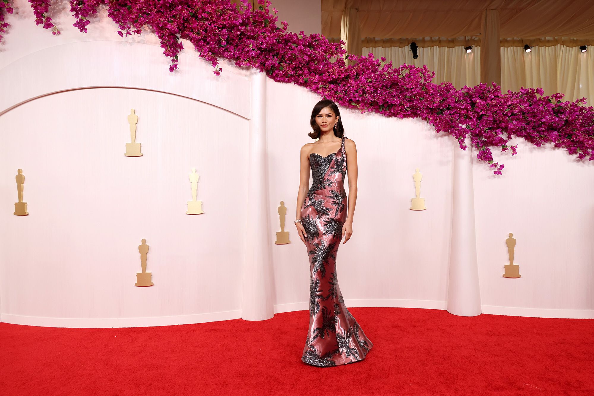 A celebrity walking the Oscars red carpet in Los Angeles wearing a classic black-tie formal look with bright flash photography and step-and-repeat backdrop