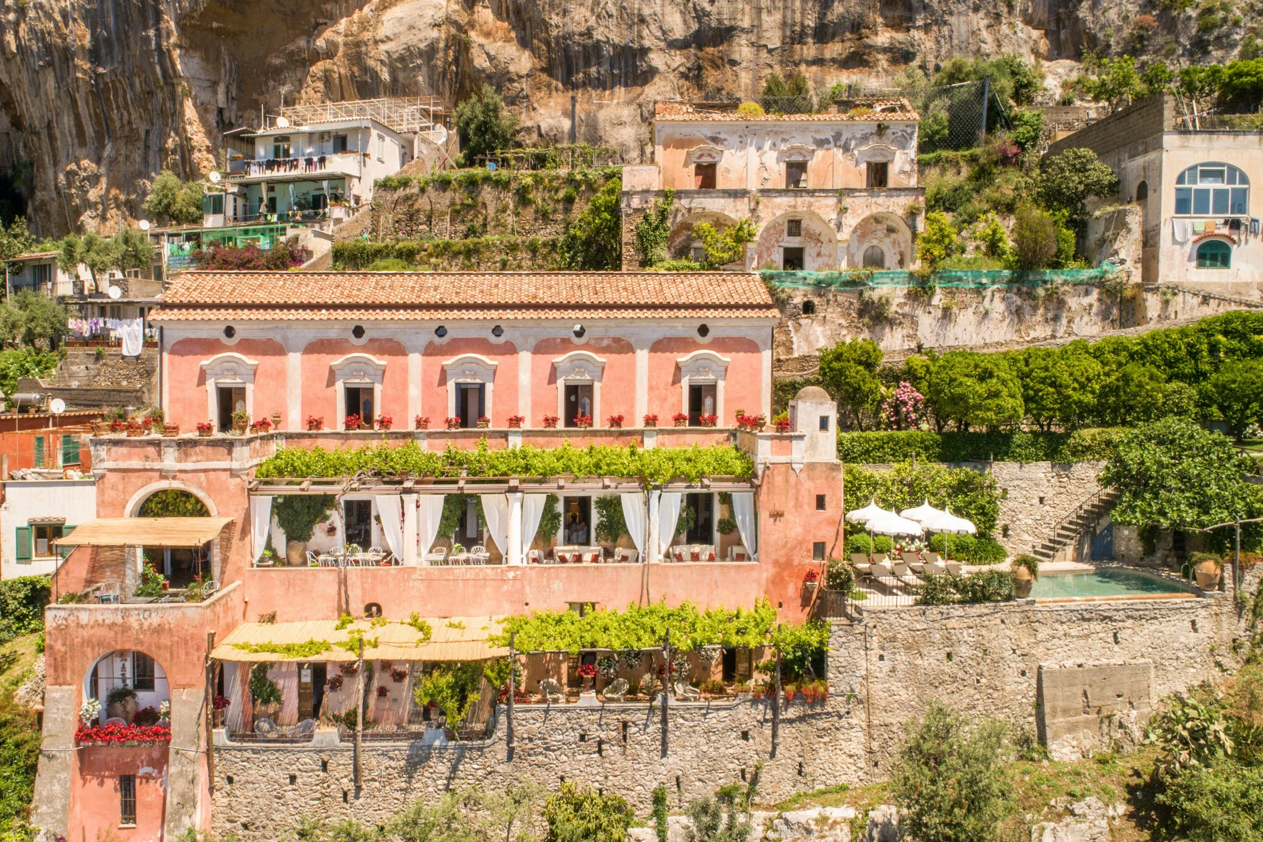 A cliffside villa on the Amalfi Coast with pastel buildings nearby and a sunlit terrace overlooking the sea