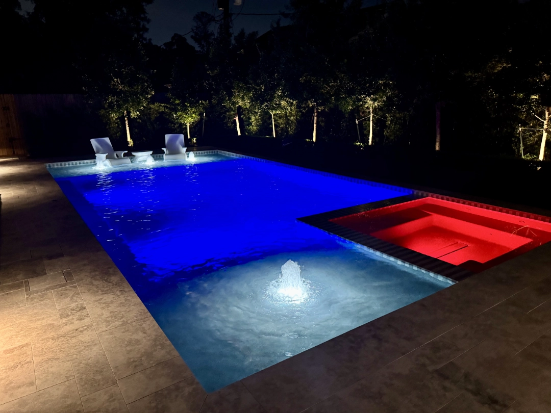 A contemporary backyard with a rectangular pool lit by underwater LEDs, simple stone coping, and subtle landscape lighting along a hedge at night