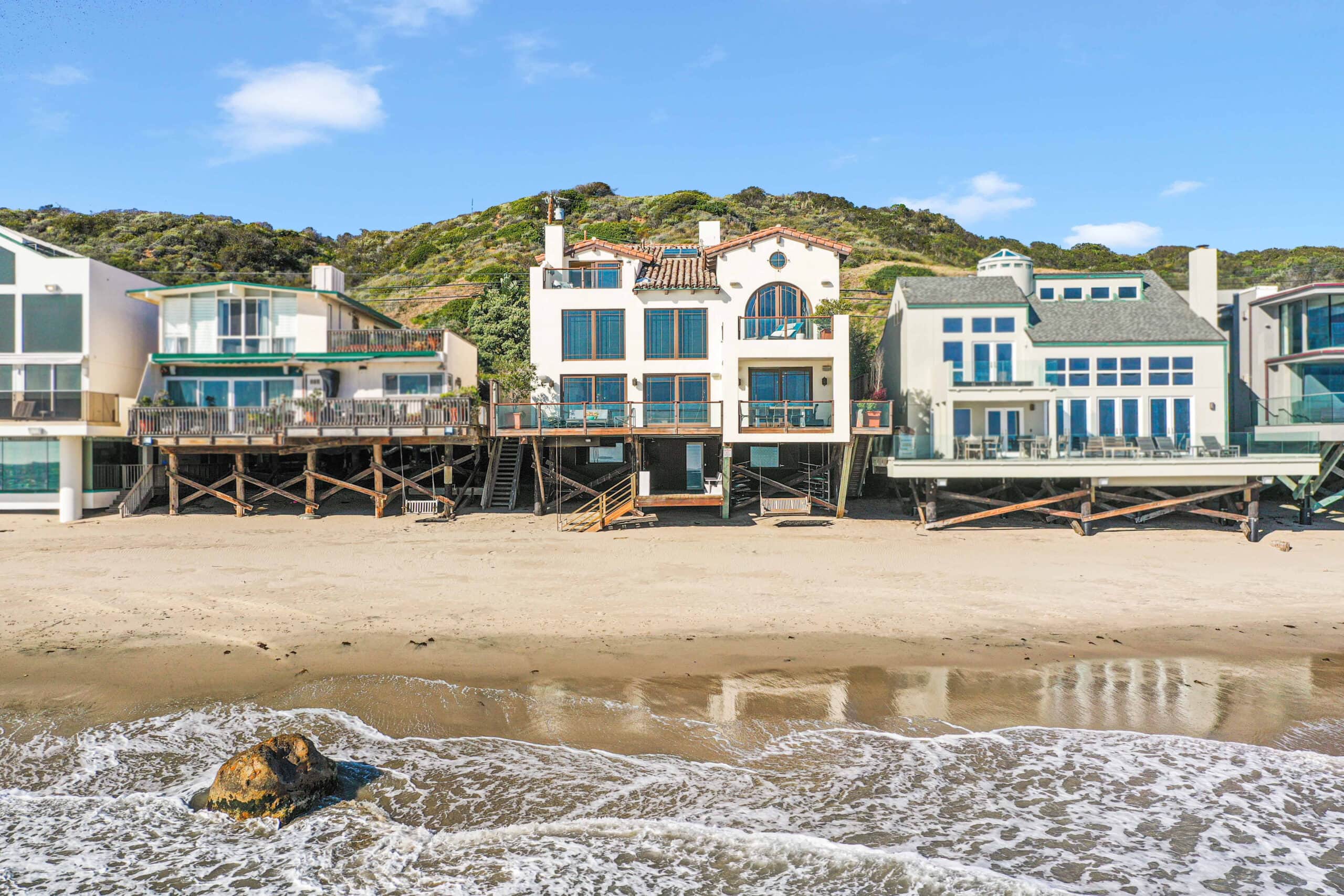 A contemporary beachfront house in Malibu with floor-to-ceiling windows, a wooden deck, and gentle waves rolling in behind it