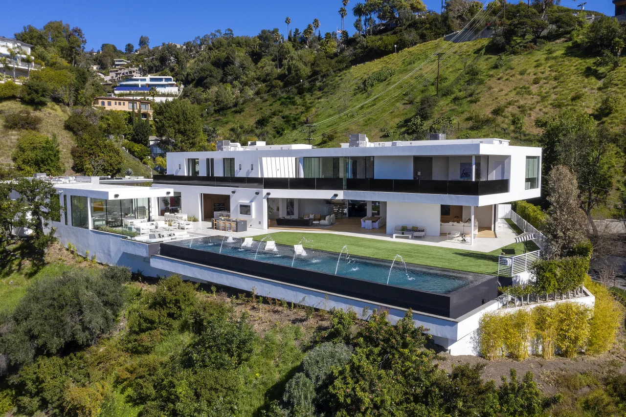 A crisp daytime photograph of a modern Beverly Hills hillside mansion with a long driveway, tall hedges, and a glass-fronted facade
