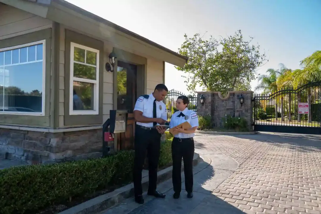 A gated Los Angeles residential street lined with hedges and privacy walls, photographed in daylight, with visible home security features