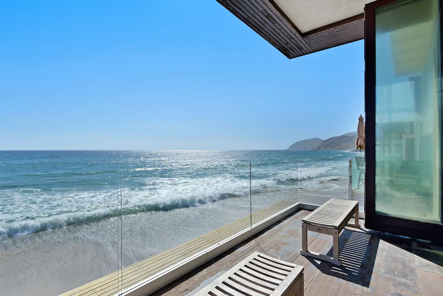 A golden-hour photograph of a modern Malibu oceanfront home with large glass doors opening to a patio facing the Pacific