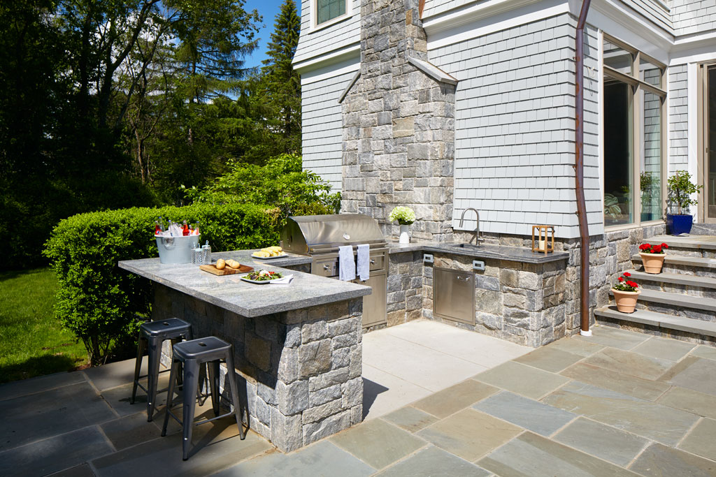 A high-end outdoor kitchen with a built-in stainless steel grill, a stone island with bar stools, and warm pendant lighting under a covered patio