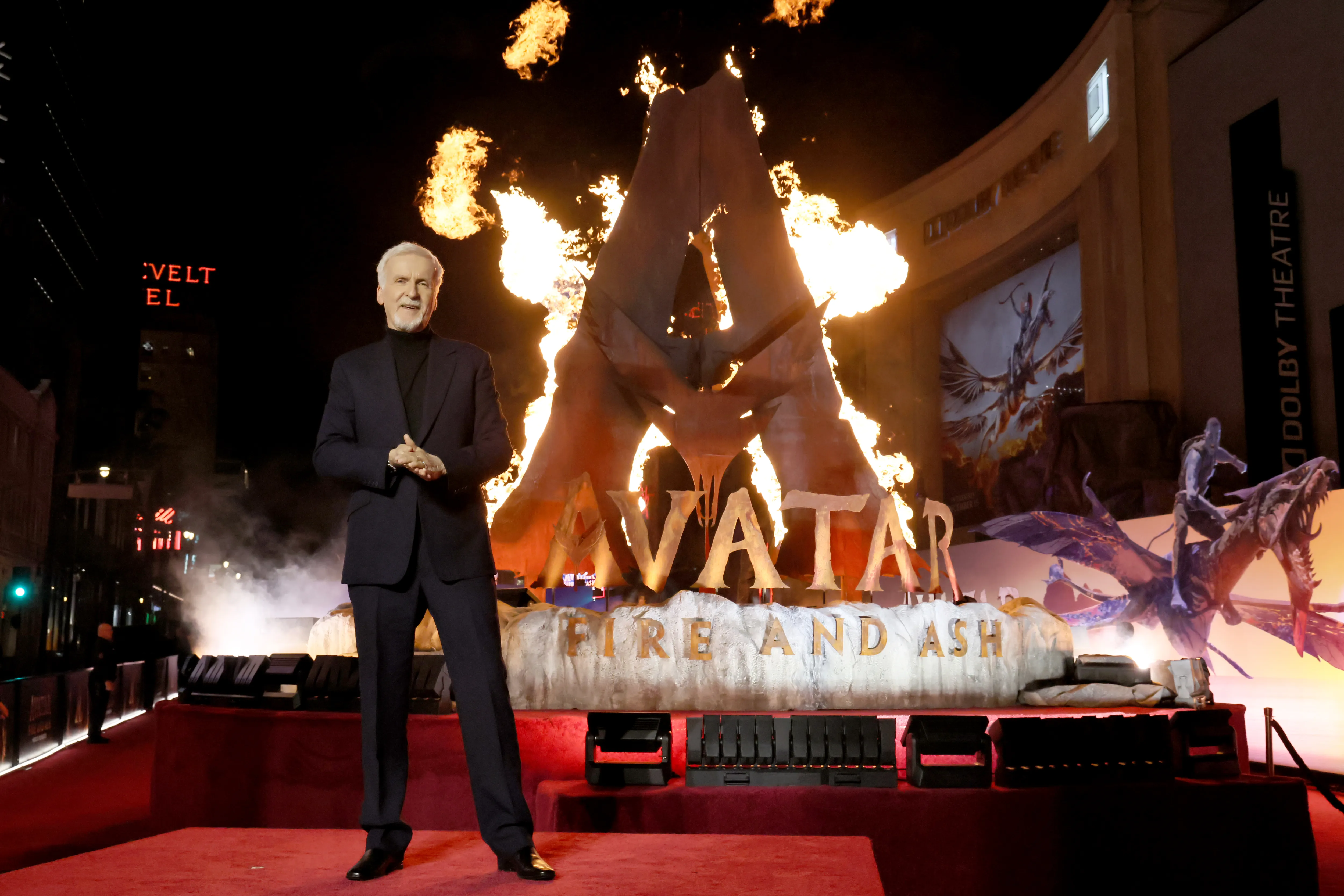 A nighttime movie premiere arrival line outside a major theater, with velvet ropes, bright lights, photographers, and guests stepping onto the carpet