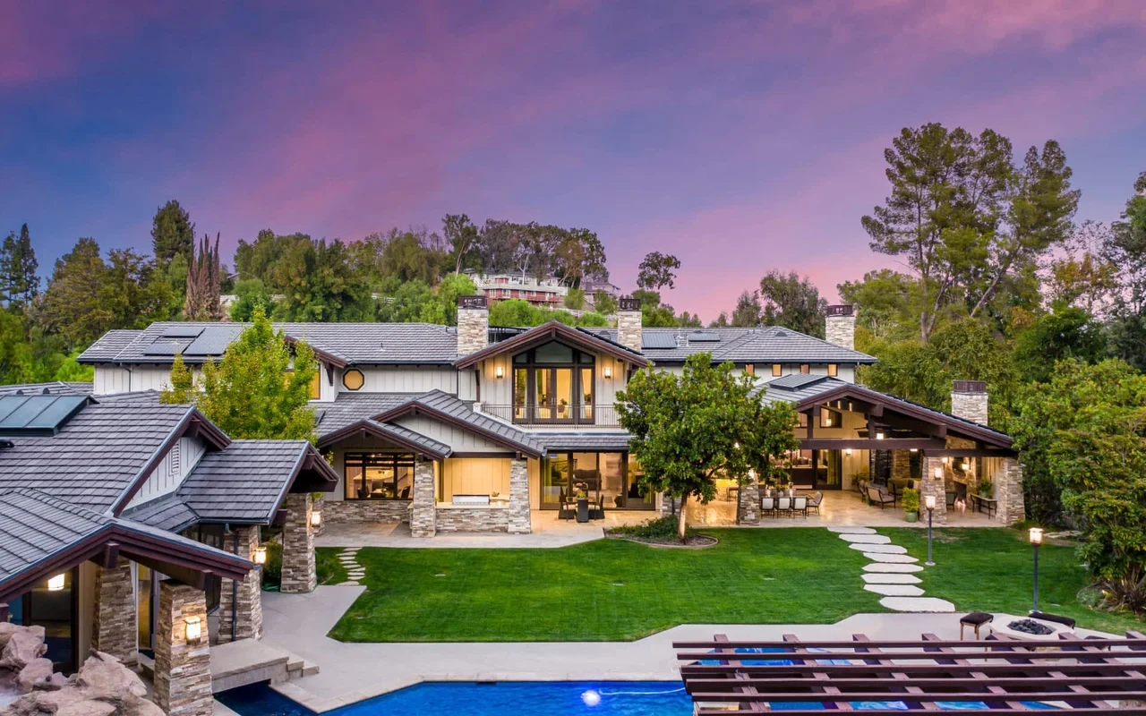 A real photograph of a modern luxury home in Calabasas with tall privacy hedges, a long driveway, and a gated front entry, photographed in the late afternoon