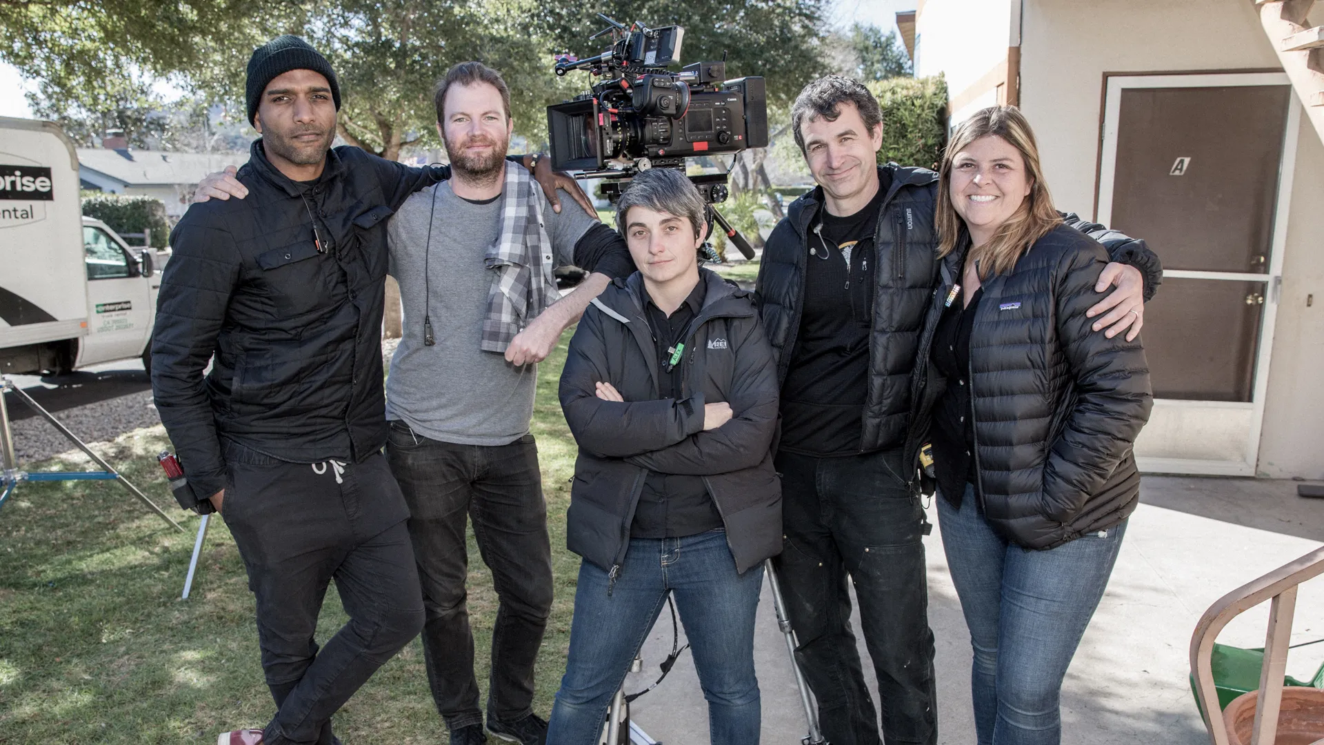 A small independent film crew setting up lights and a camera in a modest interior location while a director reviews notes