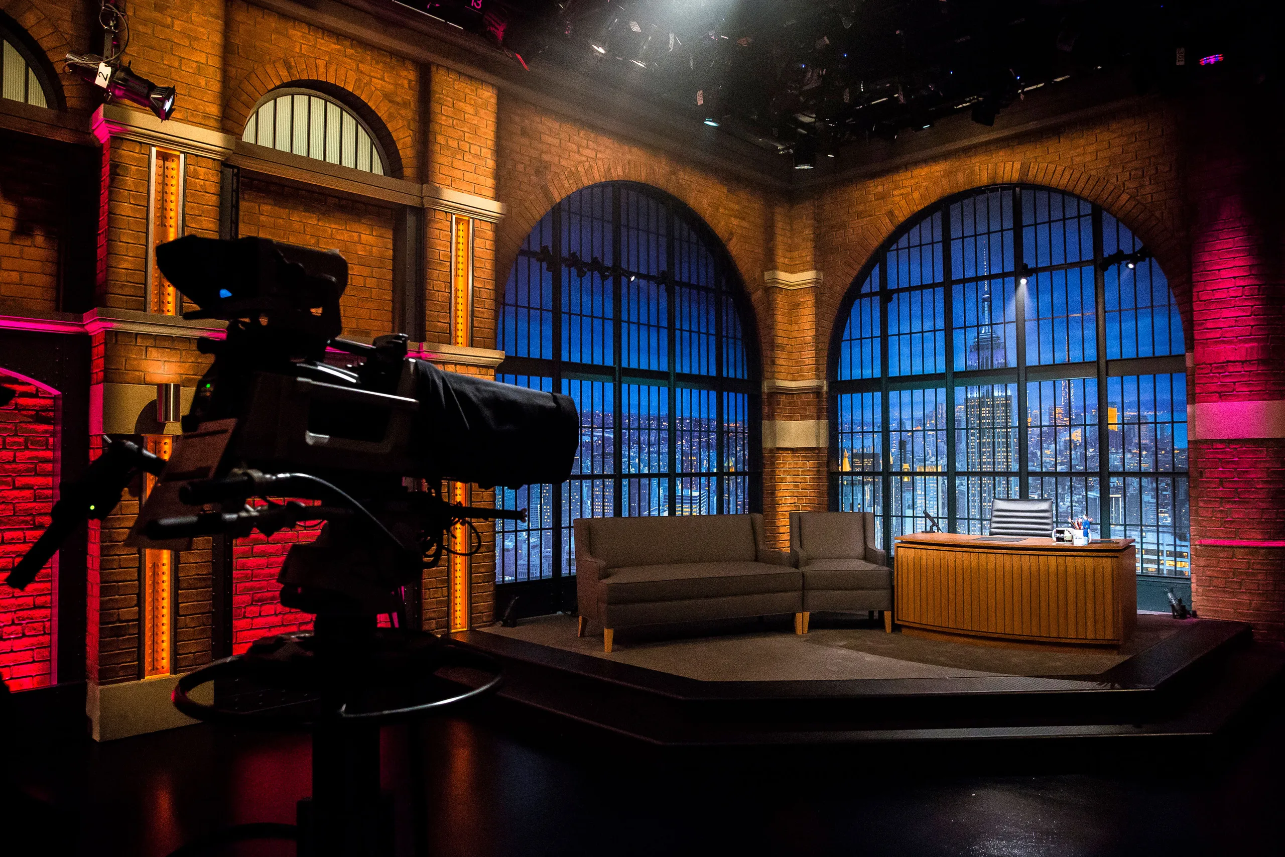 A wide shot of a late-night talk show studio with audience seating, stage lights glowing, and a host desk visible from the crowd