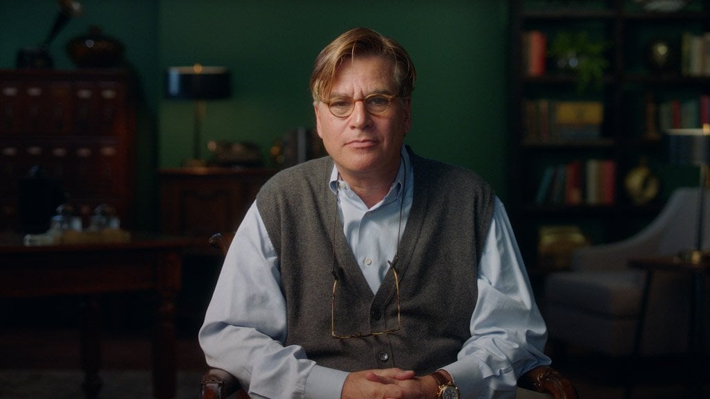 Aaron Sorkin sitting at a desk in a dark studio set, gesturing as he explains a screenwriting concept to camera