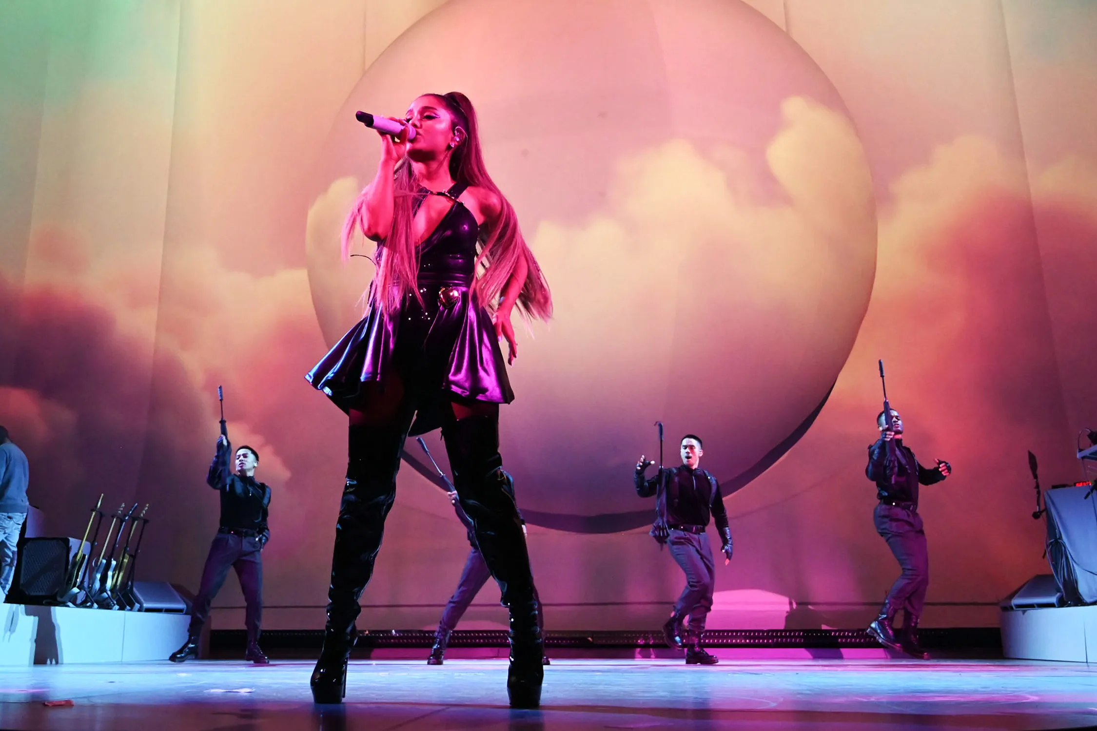Ariana Grande performing onstage during the Sweetener World Tour, holding a microphone under pink and purple stage lighting