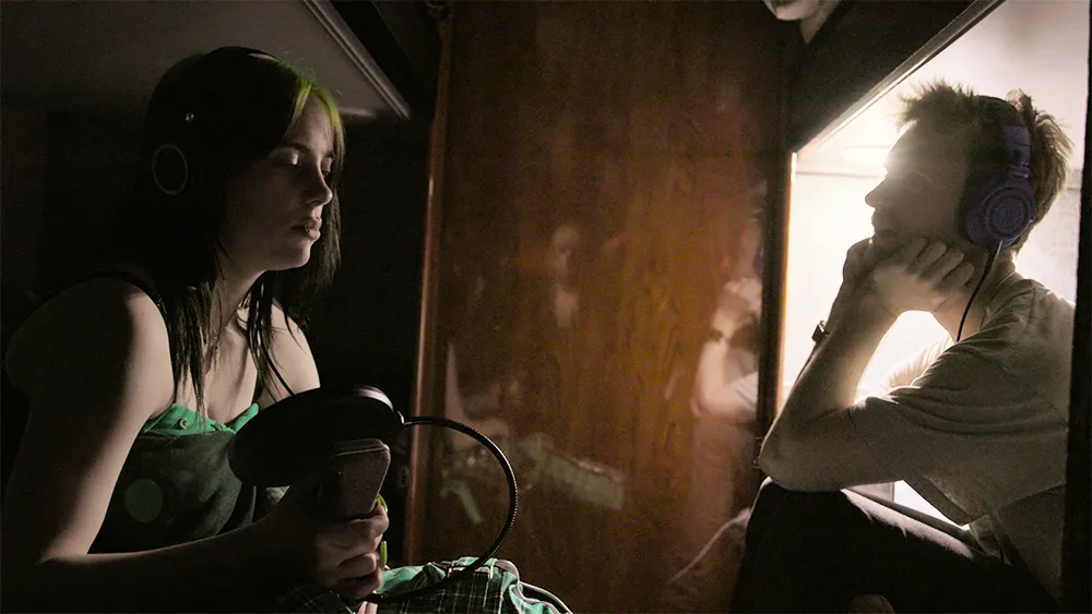 Billie Eilish sitting in a dim recording studio, wearing casual clothes and headphones around her neck, looking thoughtful between takes