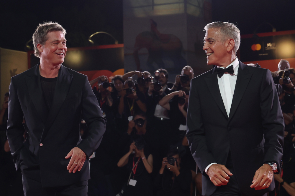 Brad Pitt standing on a film festival red carpet in a tailored suit, smiling toward photographers