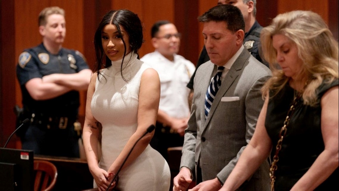 Cardi B walking outside a courthouse in a tailored outfit with security nearby as photographers take pictures.