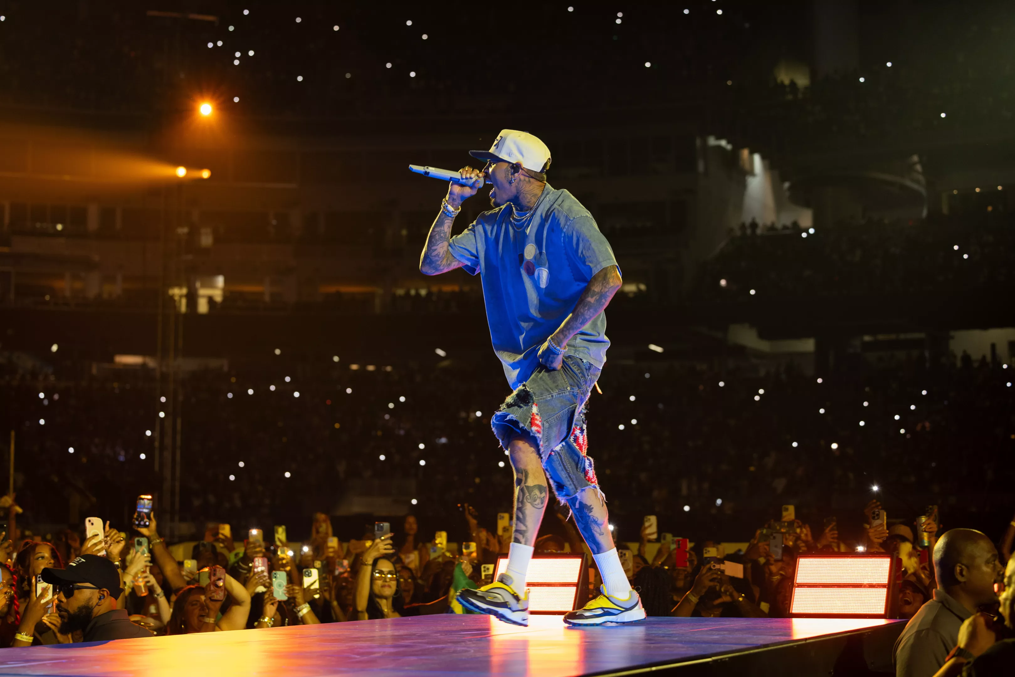 Chris Brown performing on stage under concert lighting, holding a microphone mid-song, with fans visible beyond the barricade