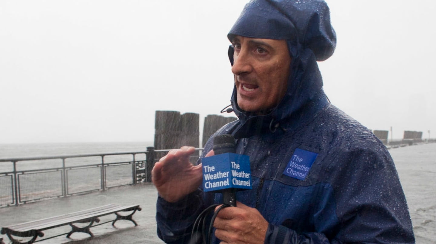 Jim Cantore holding a microphone outdoors during severe weather coverage, wearing a rain jacket as strong wind and rain hit him during a live report