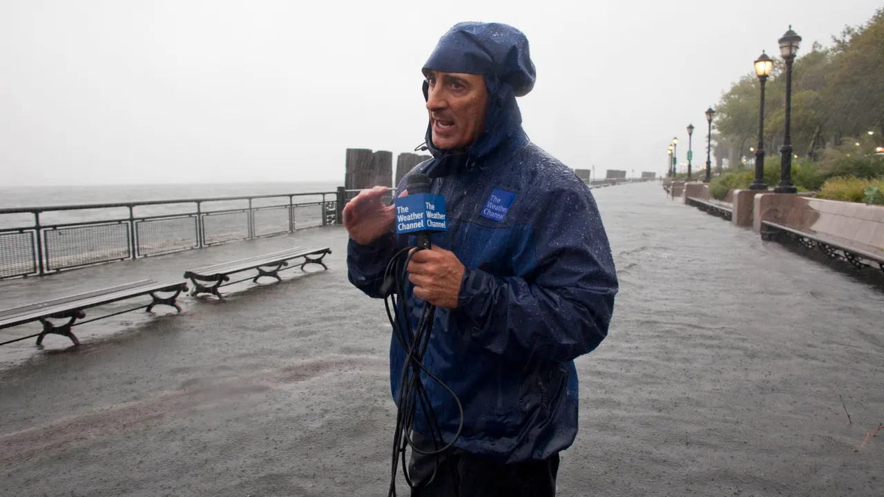 Jim Cantore reporting live during hurricane conditions near a coastline, with palm trees bending in the wind and heavy rain blowing across the street