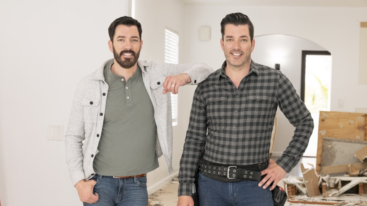 Jonathan Scott wearing work boots and holding renovation plans inside a partially updated living room with exposed beams