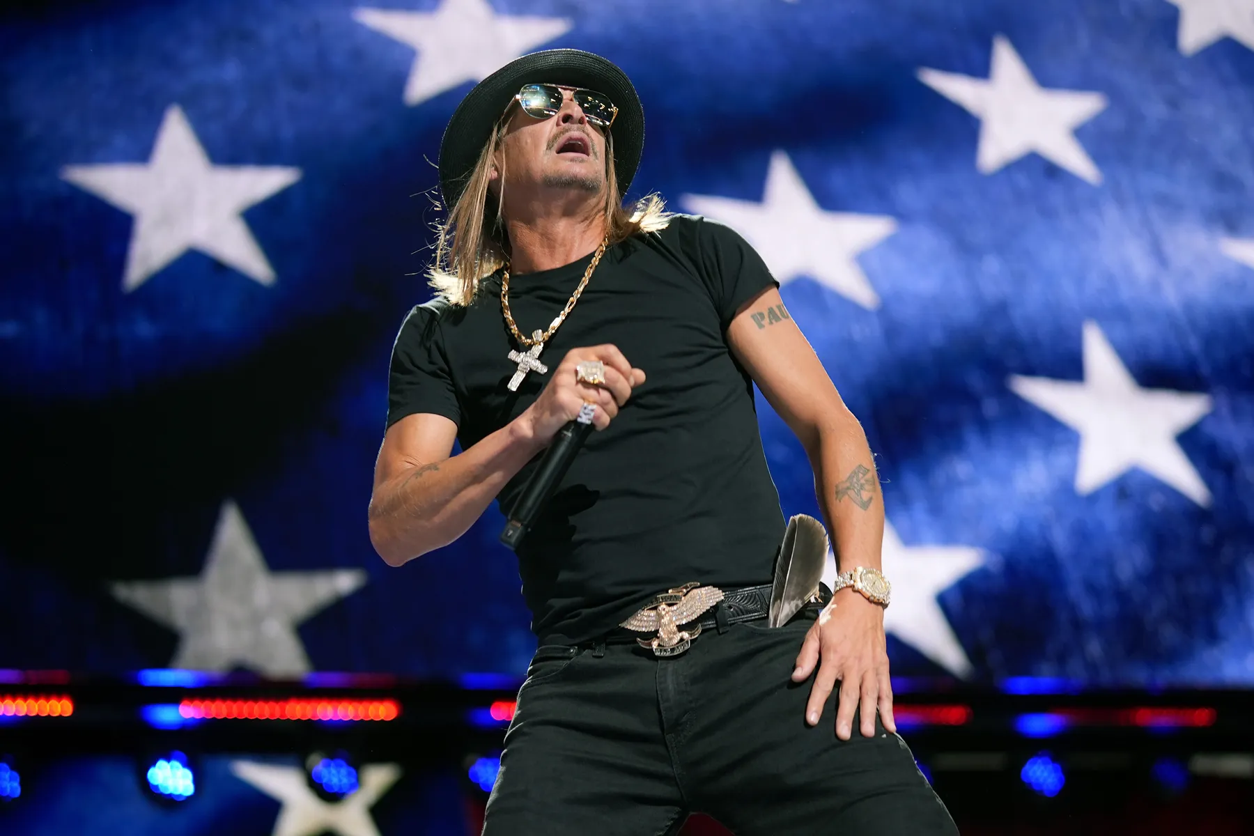 Kid Rock performing onstage under bright concert lights, holding a microphone and facing the crowd