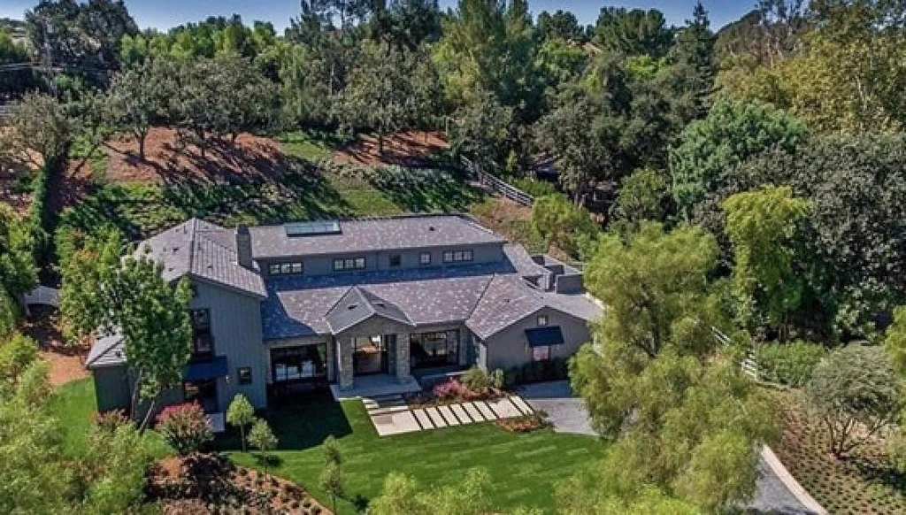 Kris Jenner stepping outside a modern Hidden Hills style home with a long driveway, tall hedges, and a minimalist exterior