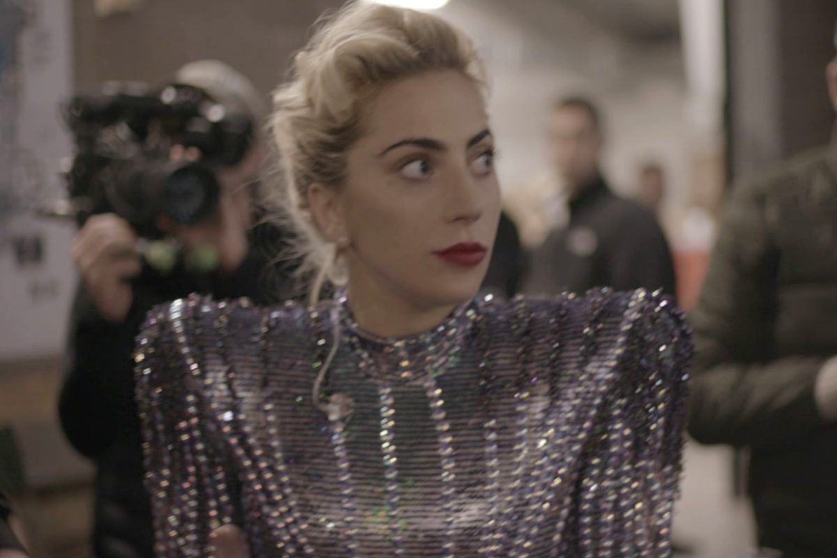 Lady Gaga backstage in a dressing room, wearing a robe with her hair pinned up, looking tired but focused before a performance