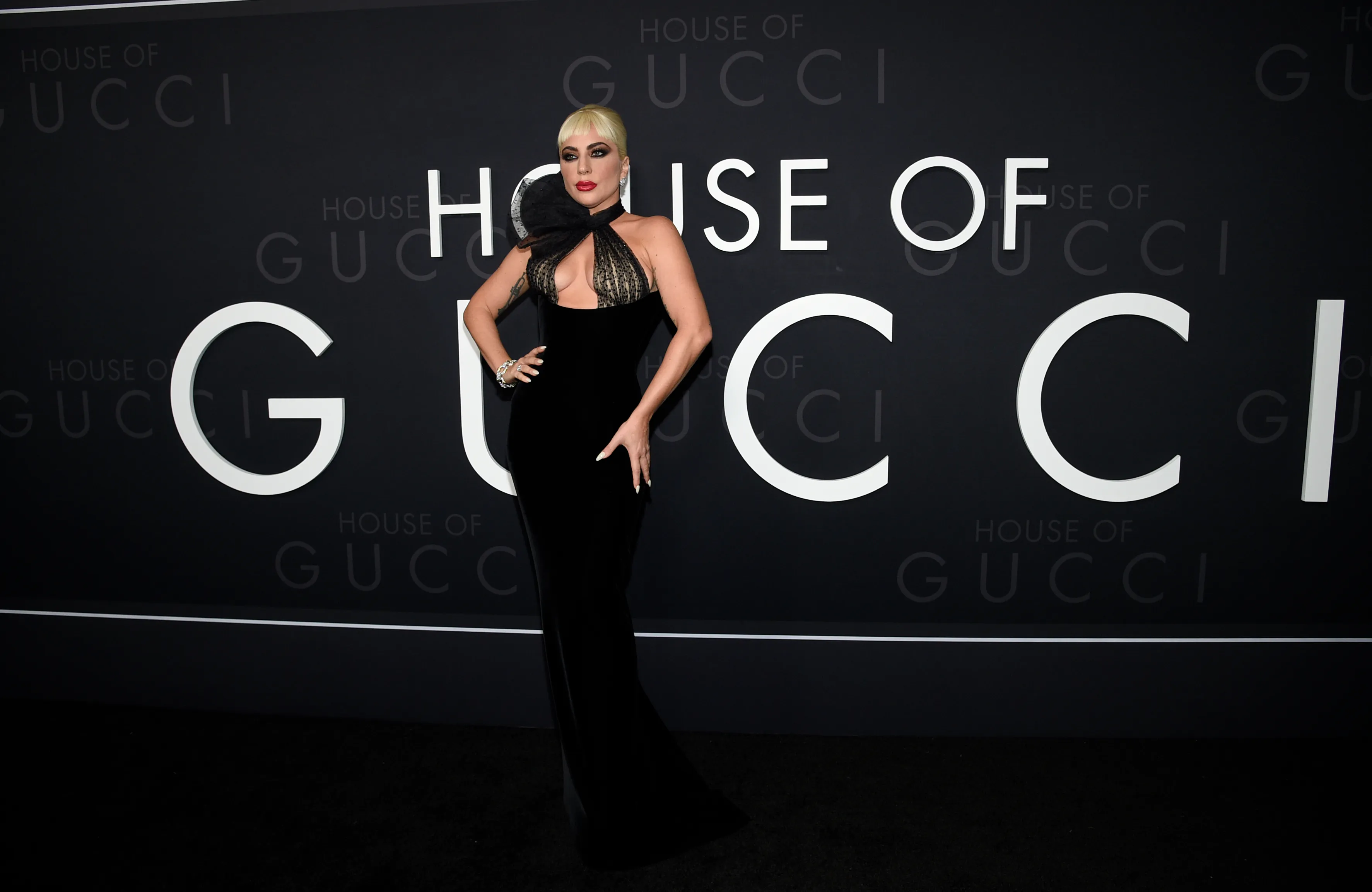Lady Gaga walking a red carpet in a dramatic, statement-making gown with high-glam hair and makeup, turning toward cameras as flashes go off