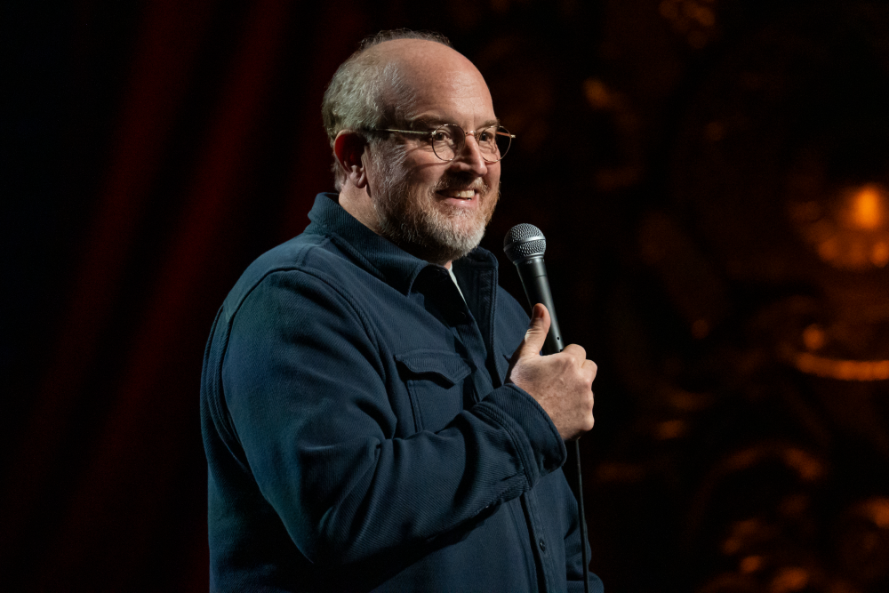 Louis C.K. performing standup onstage under warm spotlight lighting with a handheld microphone