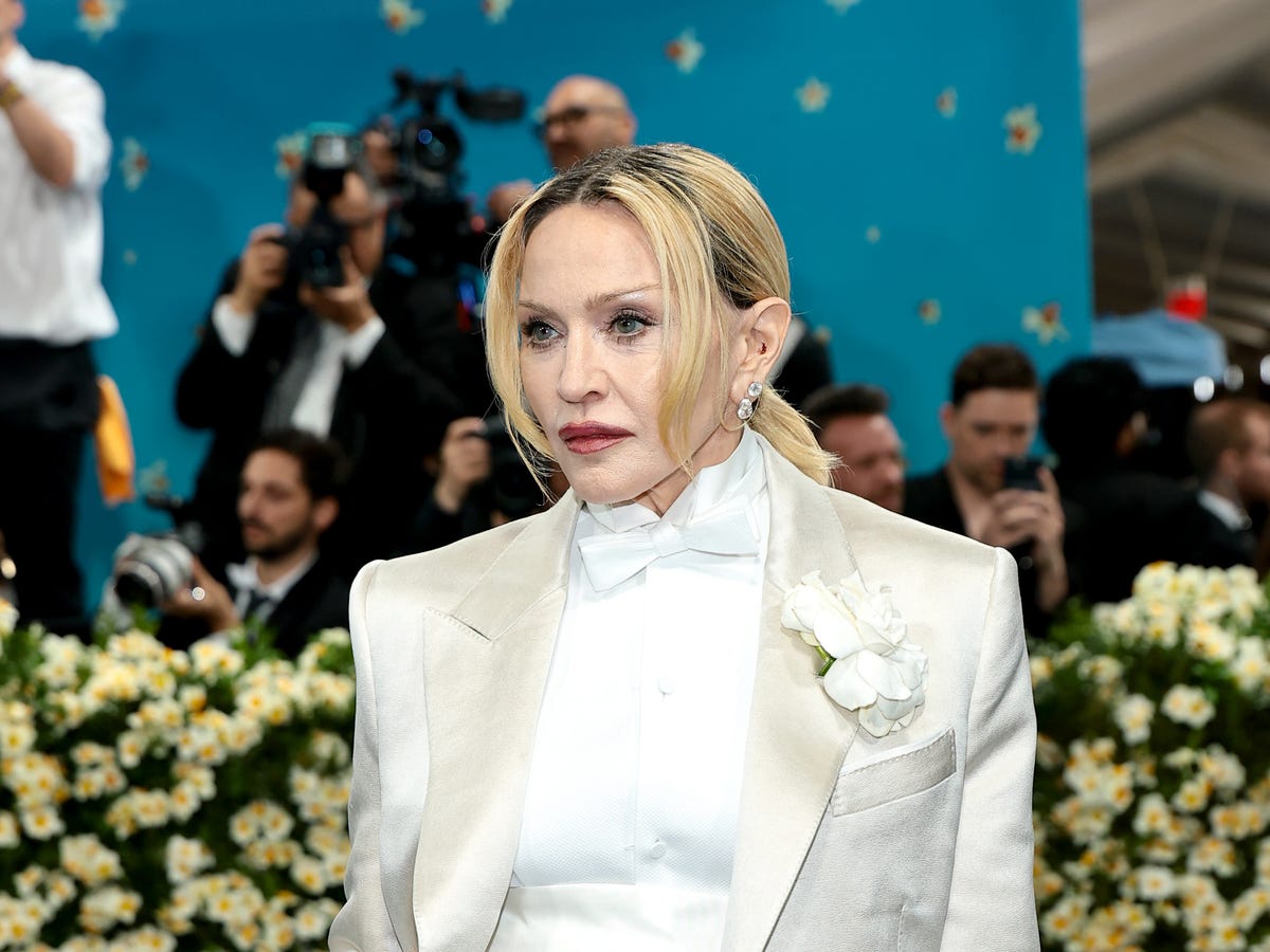 Madonna posing for photographers on a red carpet in a dramatic fashion look, with event lighting and a step-and-repeat backdrop behind her.