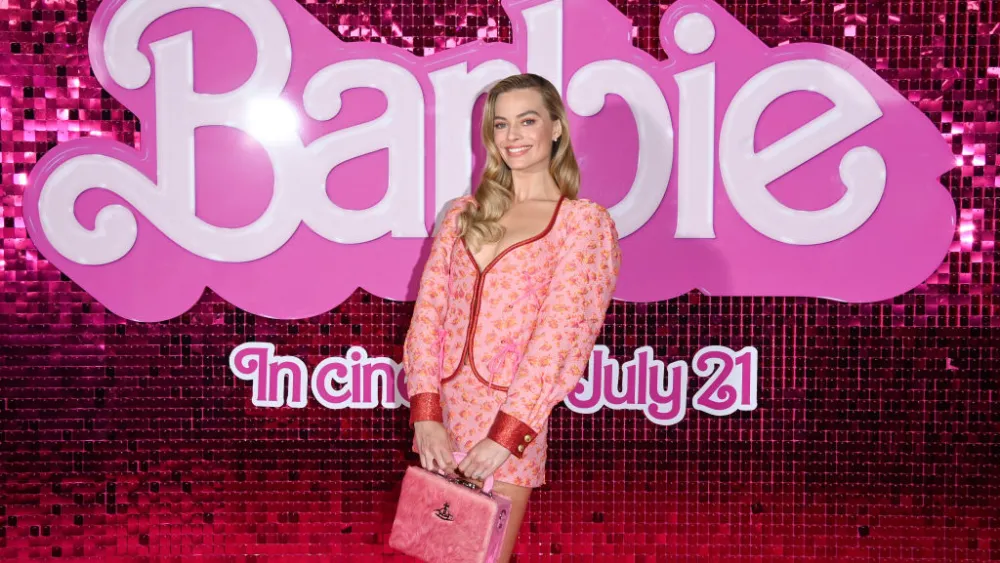 Margot Robbie arriving for a press event in a bright, Barbie-inspired ensemble with coordinated accessories, smiling toward photographers under event lighting