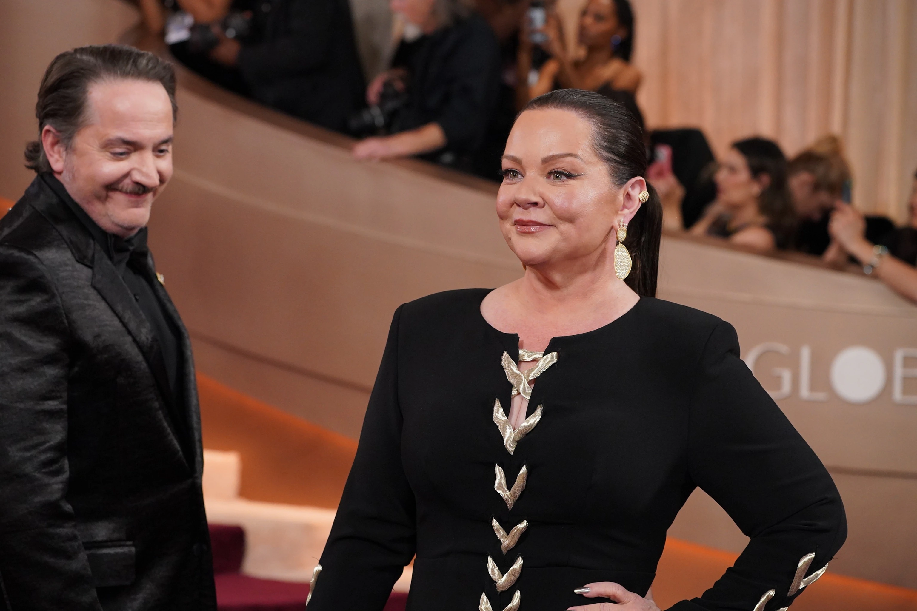 Melissa McCarthy on an awards show red carpet in a draped, elegant gown with a flattering neckline and a smooth, column-like silhouette, smiling toward photographers