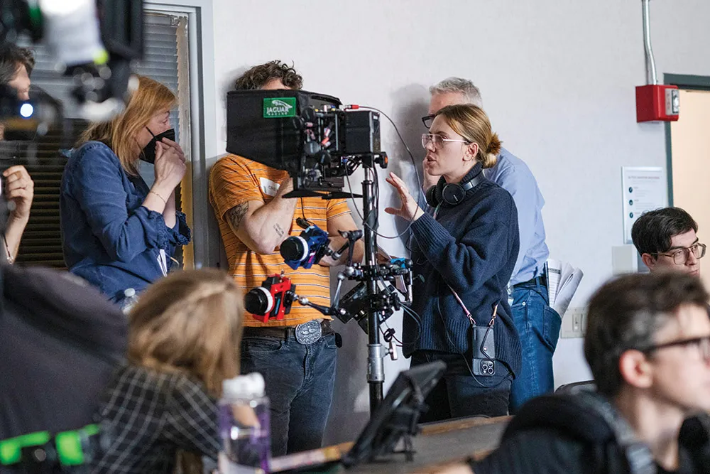 Scarlett Johansson on a film set wearing a headset, standing near a camera rig while looking toward actors off-frame