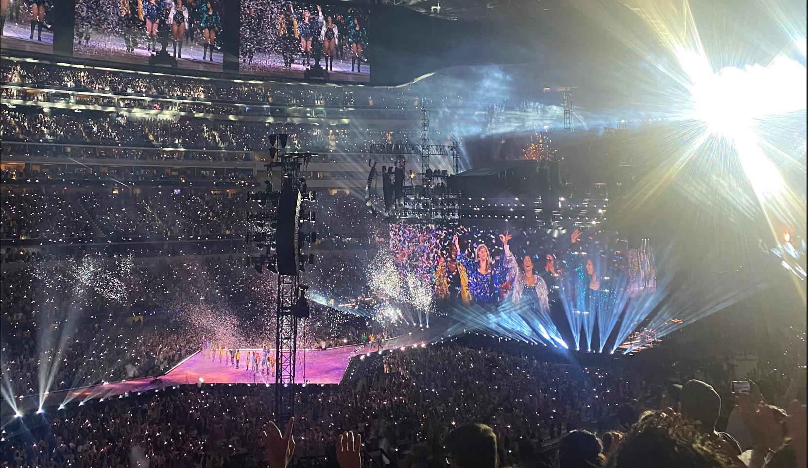 Taylor Swift performing onstage in a stadium concert setting, wearing a stage outfit with dramatic lighting and fans visible in the background.