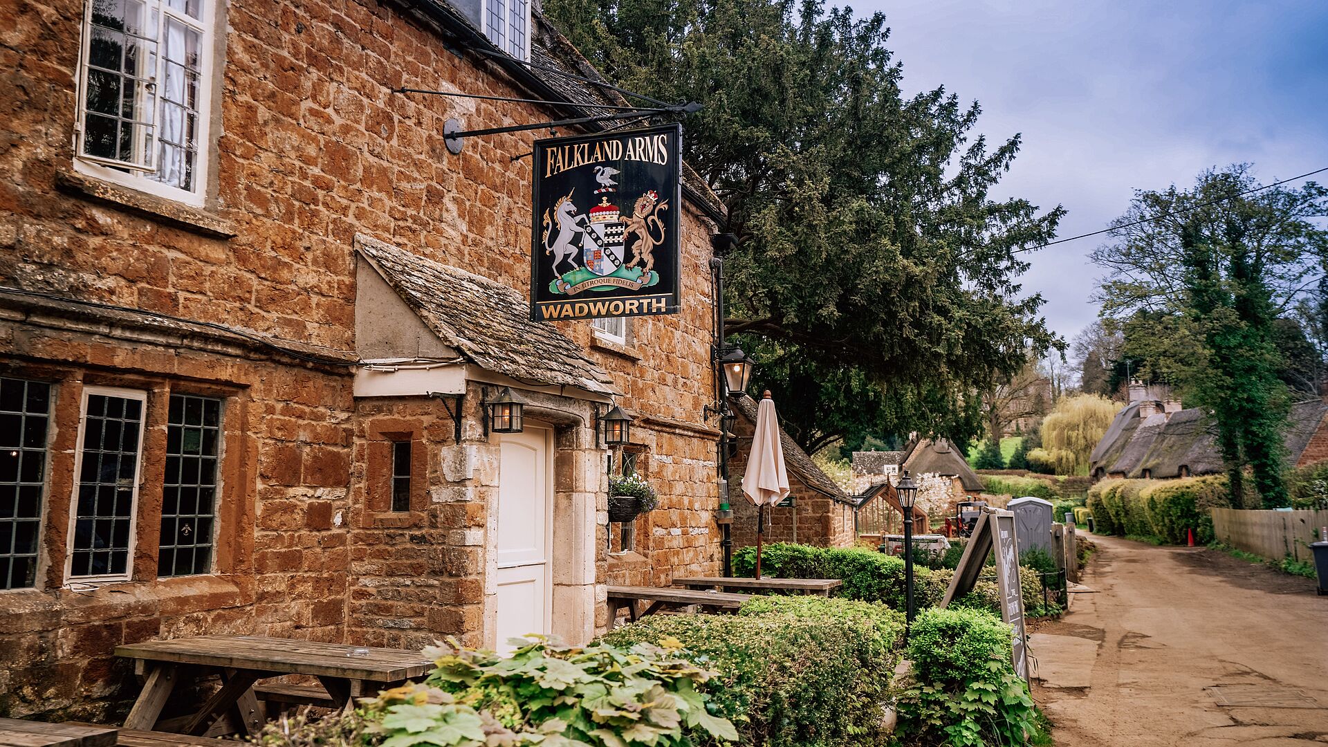 The exterior of The Falkland Arms, a historic pub and boutique inn in Great Tew, Oxfordshire, with traditional stone architecture and signage visible in daylight