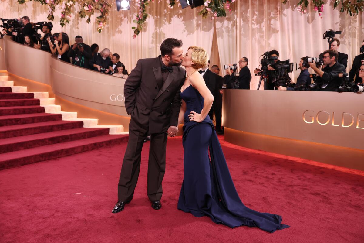 Two celebrities standing on a Golden Globes style red carpet with step-and-repeat logos behind them, posing with a formal posture as photographers call out
