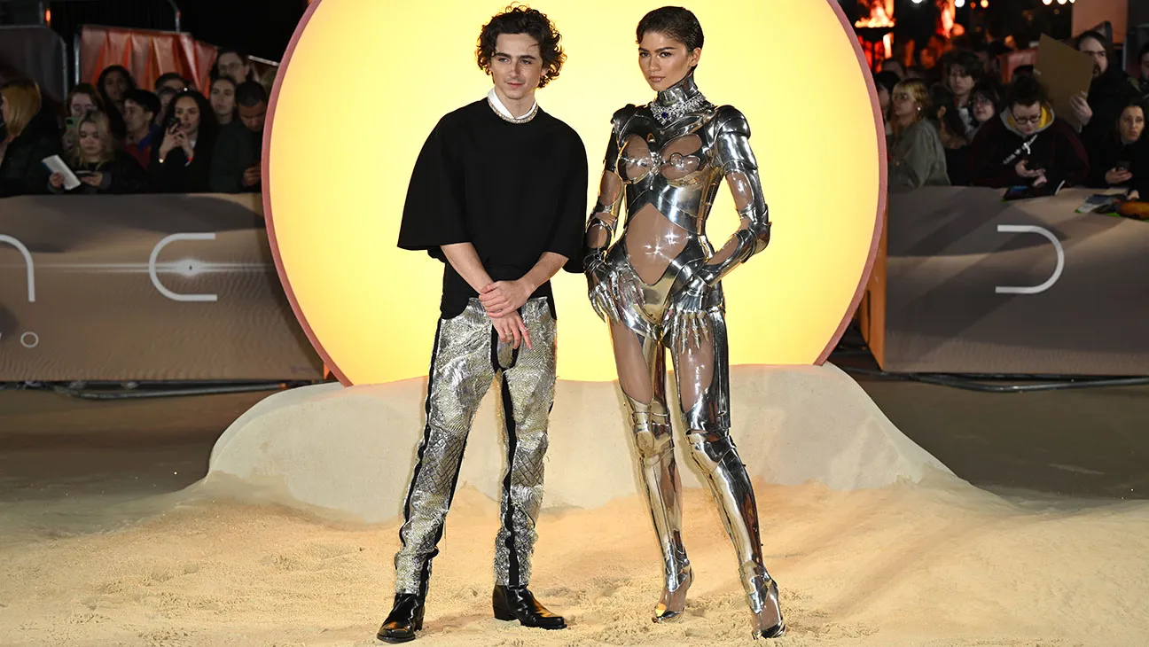 Zendaya posing on a red carpet in a sleek, high-fashion gown with sculptural details and dramatic lighting reflecting off the fabric, photographers visible behind her